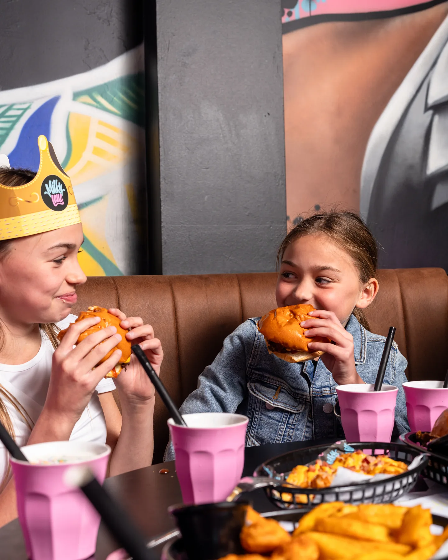 Two kids at a Milky Lane booth · one biting into a Milky Lane burger, thickshake with straw on the table